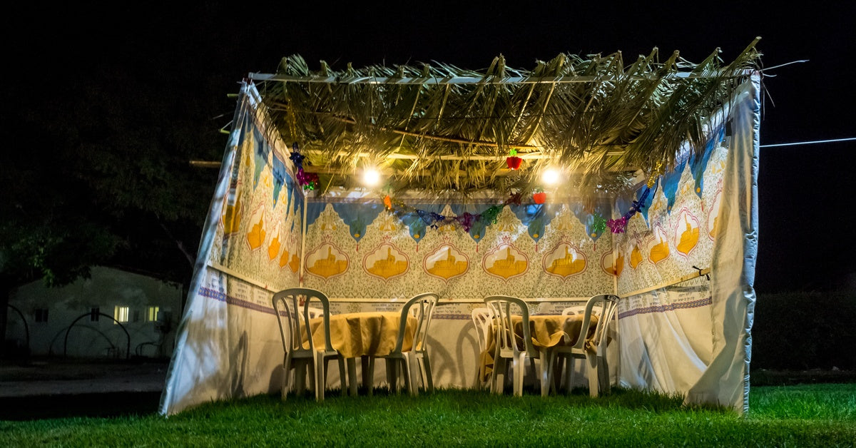 A small sukkah at night, decorated with homemade décor and two small tables in anticipation of Sukkot dinner.