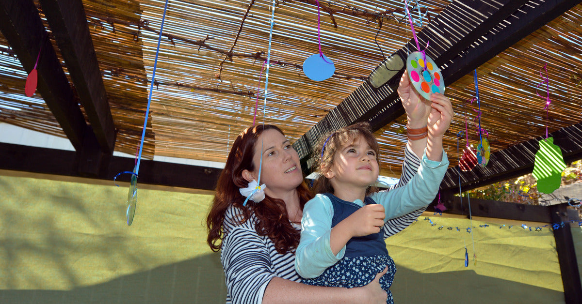Mother and child hanging decorations from bamboo schach roof – family preparing sukkah interior for Sukkot 2025.